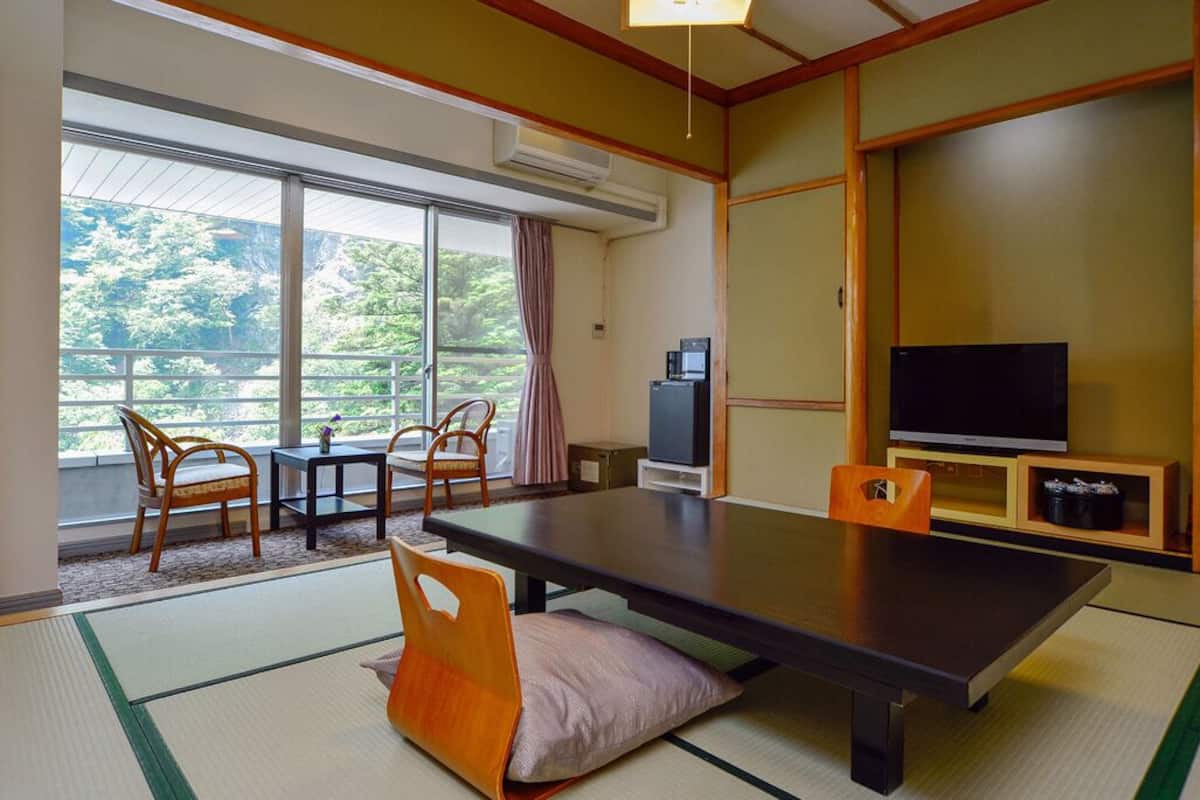 Une pièce de style japonais traditionnel, avec un sol recouvert de tatamis, une table basse, des chaises, un téléviseur et une vue sur la verdure à l’extérieur.