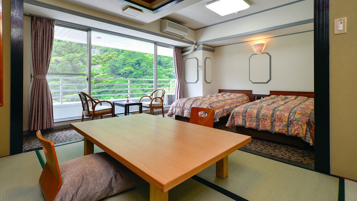 Une chambre d’hôtel avec un sol recouvert de tatamis, une table en bois, deux lits avec une literie à motifs floraux, une vue sur la verdure et un climatiseur fixé au mur.