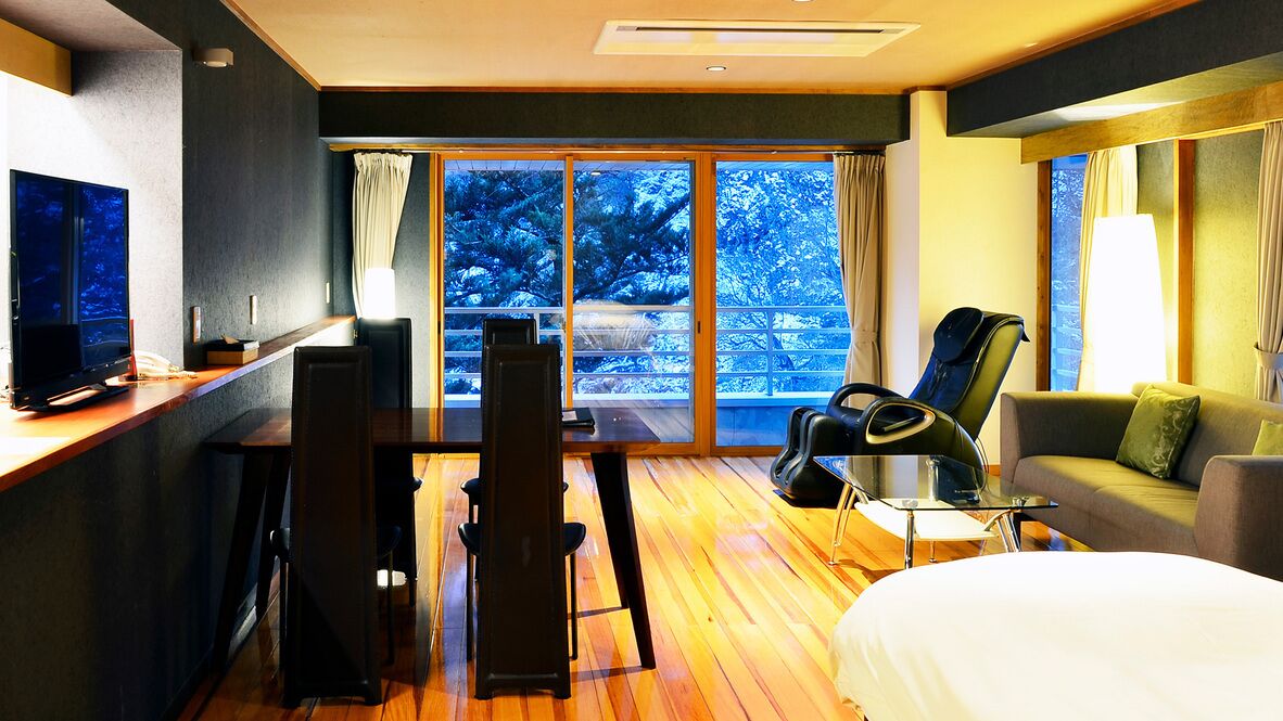 A hotel room with a bed, a desk with a computer, a chair, a sofa, and a large window with a view of trees.