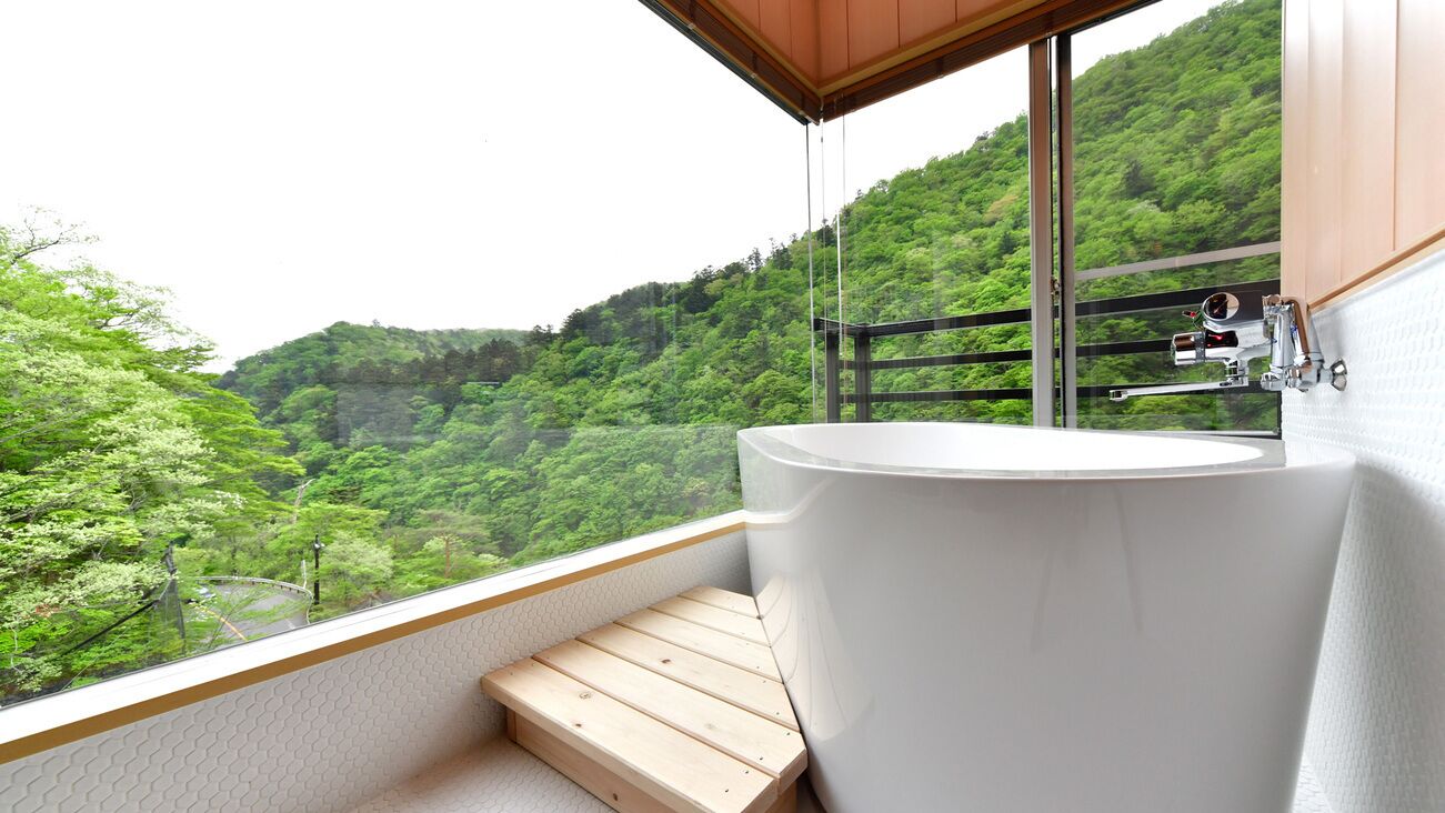 A bathroom with a large bathtub, a wooden bench, and a view of a lush green hillside.