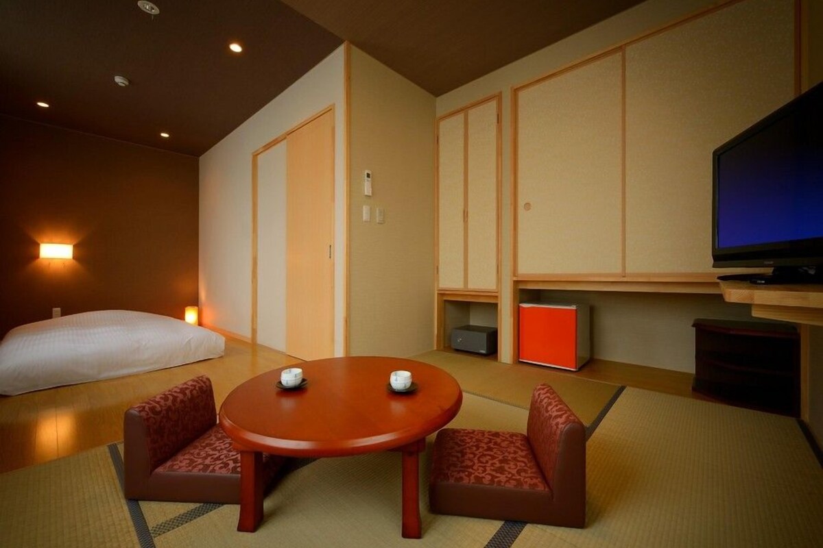 Una habitación de hotel con una cama, un televisor, una mesita con dos tazas y dos sillones con reposabrazos.