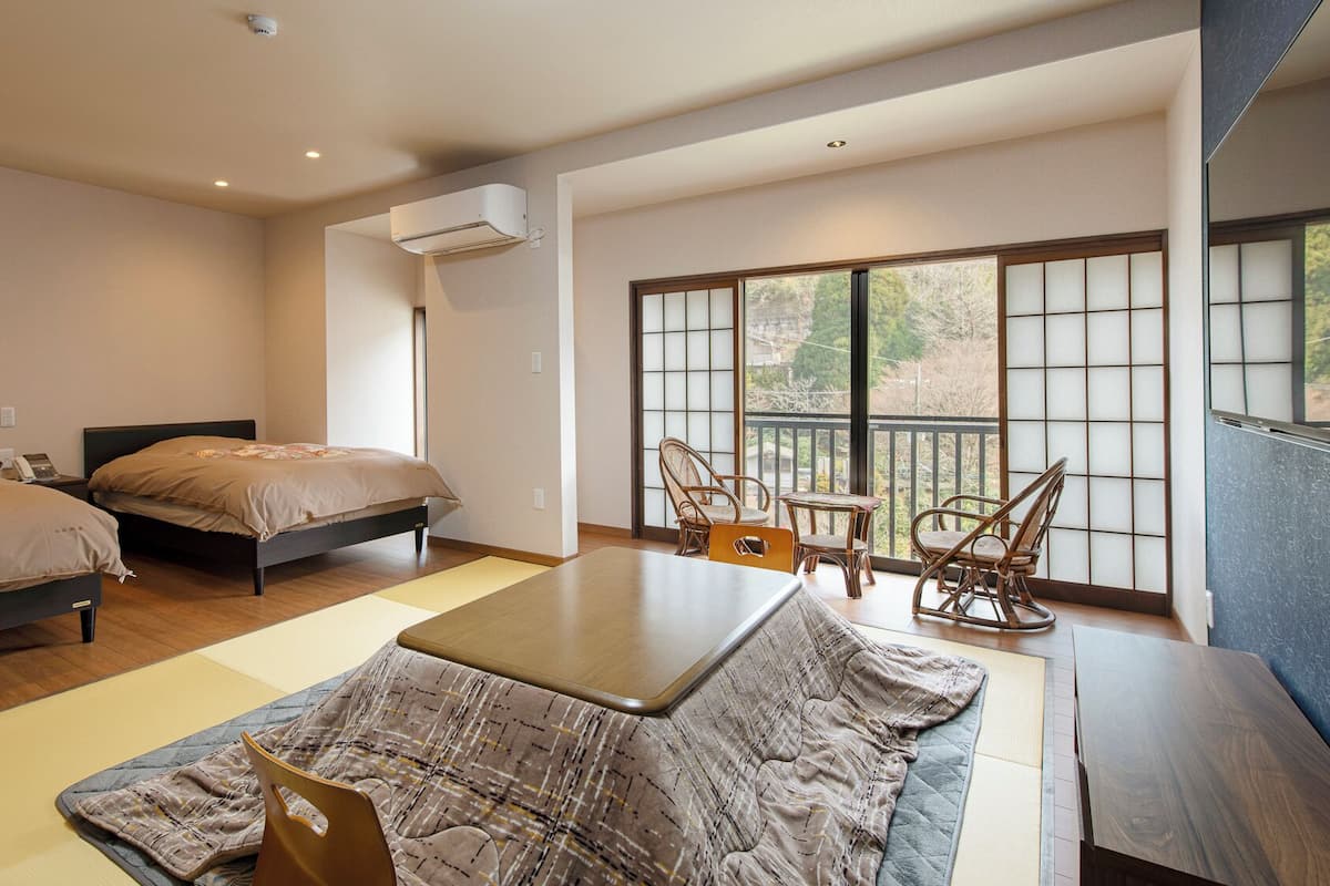 A hotel room with two beds, a dining table, chairs, a view through a large window, and an air conditioning unit.