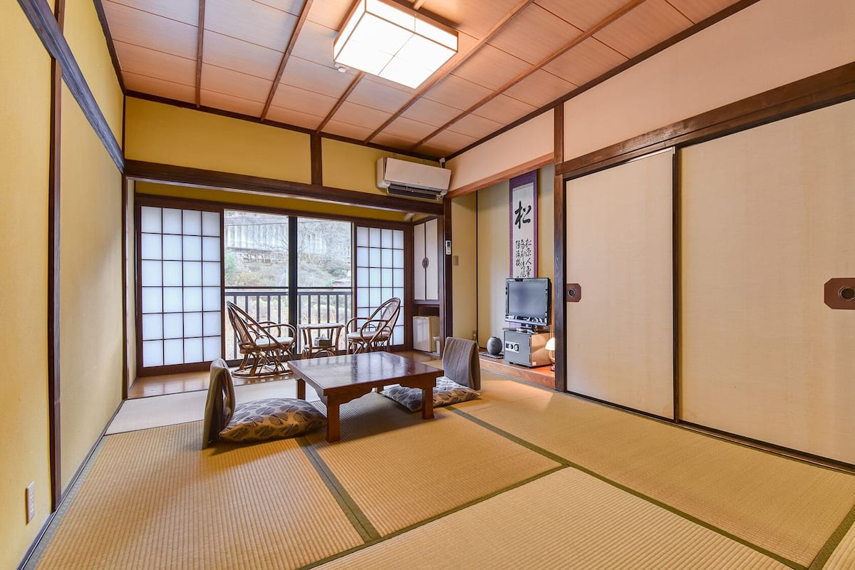 A traditional Japanese room with tatami flooring, a low table with cushions, and a television.