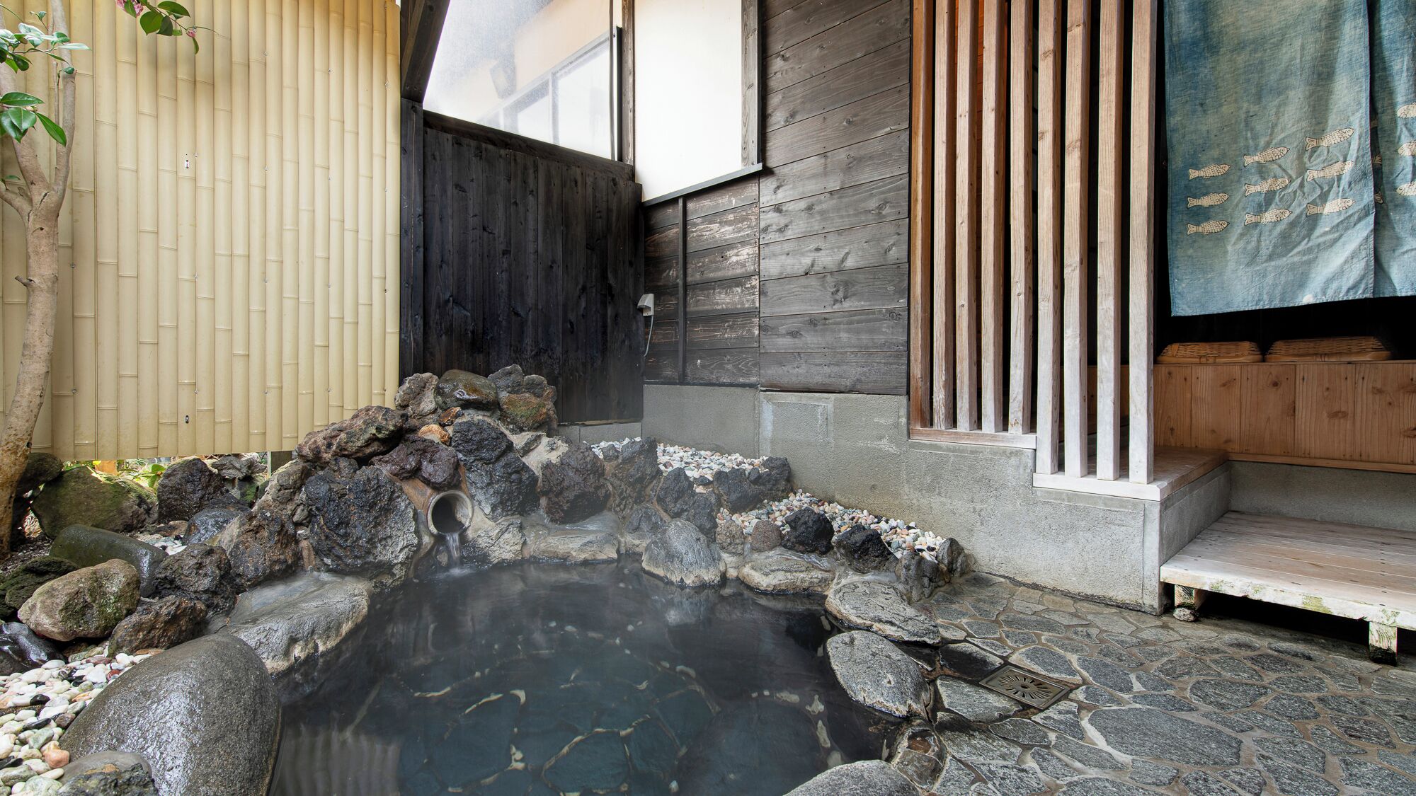A traditional Japanese onsen with a stone hot spring, wooden benches, and a bamboo fence.