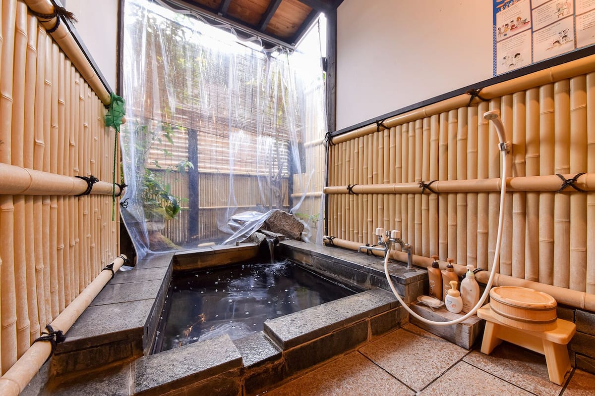 A traditional Japanese onsen with bamboo walls, a stone hot spring, and a wooden washbasin.