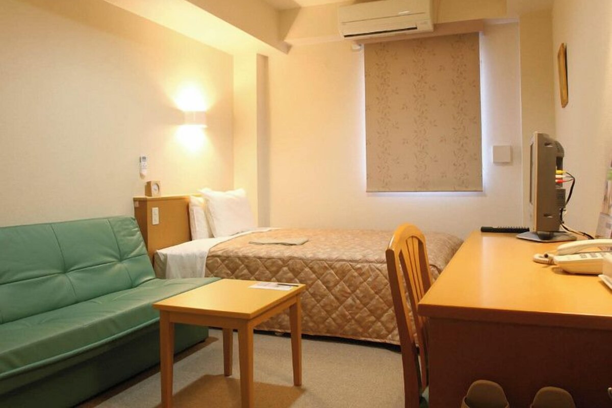 A small, cozy hotel room with a green sofa, a single bed, a wooden desk with a computer, and a window with a view.