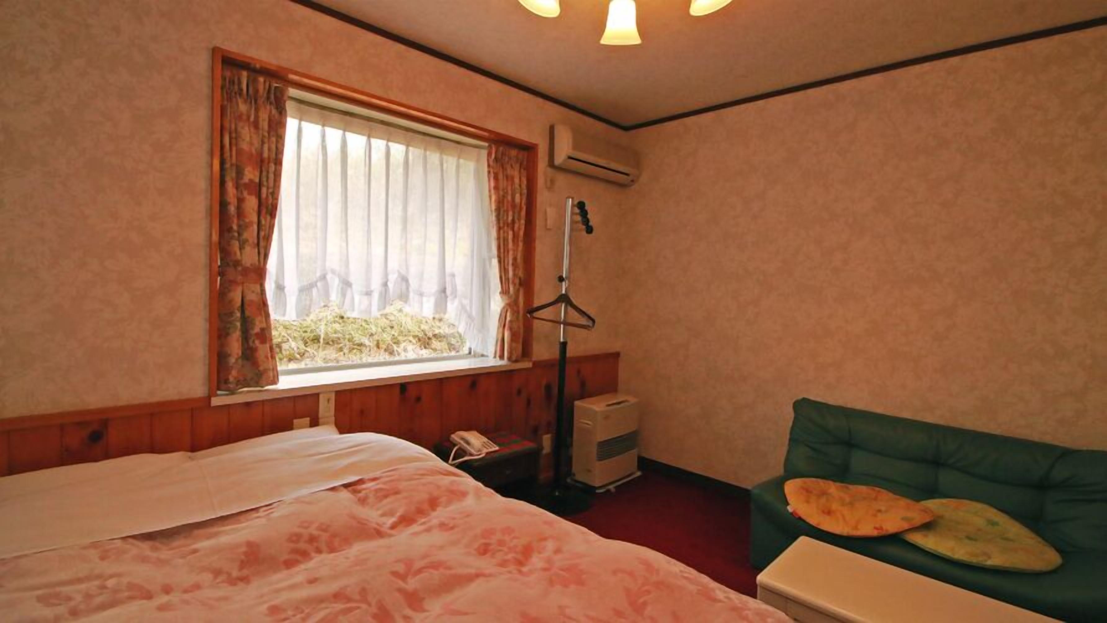 Habitación de hotel con cama, ventana con cortinas, aire acondicionado, sofá verde y una mesita con un almohadón.