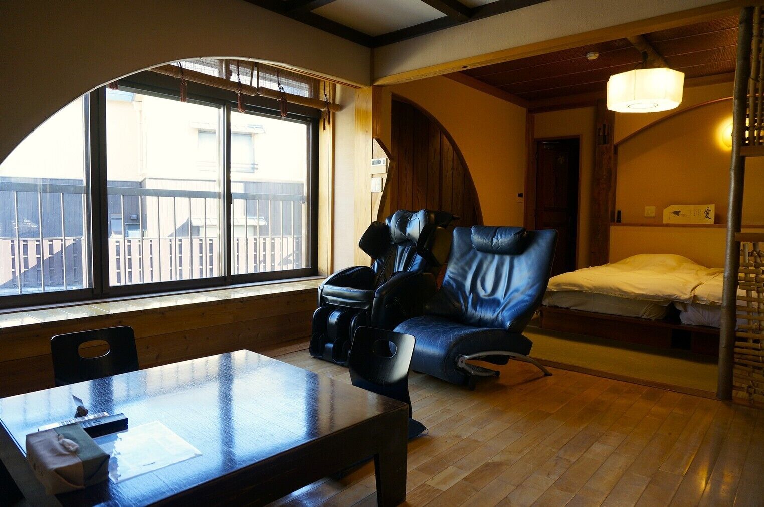 Een kamer met een houten tafel, massagestoelen, een bed en een raam met uitzicht op gebouwen.