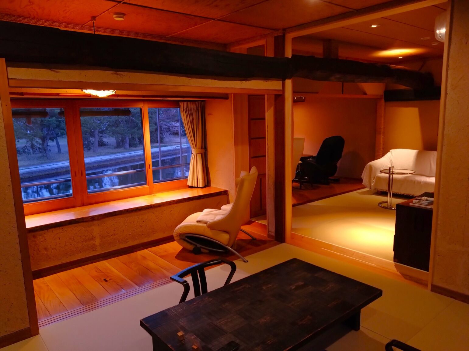 Een kamer met houten vloer, een raam met gordijnen, een witte stoel, een zwarte stoel, een witte bank en een kleine tafel.