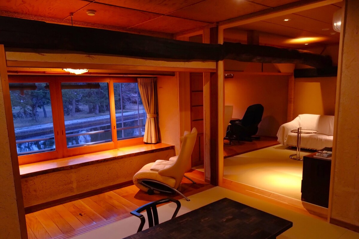 A room with a wooden floor, a window with curtains, a white chair, a black chair, a small table, and a white sofa.