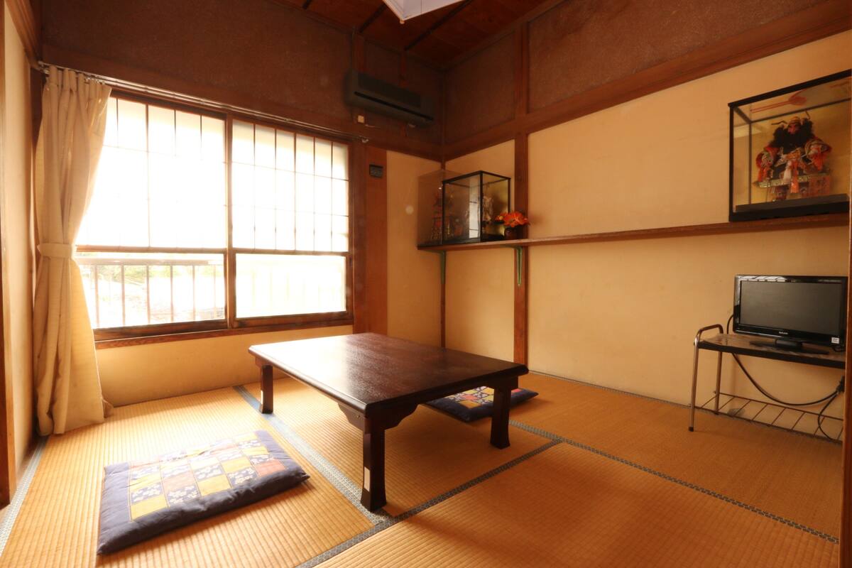 Una habitación japonesa tradicional con suelo de tatami, una mesa de madera baja, un televisor sobre un soporte y una ventana con cortinas.