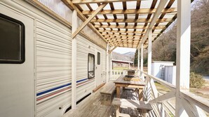 Um alpendre coberto com uma mesa de madeira e bancos, uma motorhome branca com porta deslizante, e vista da paisagem ao redor.