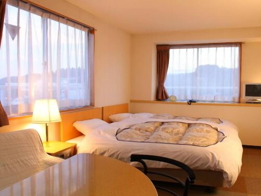 A hotel room with a bed, a desk, a chair, and two windows with curtains.