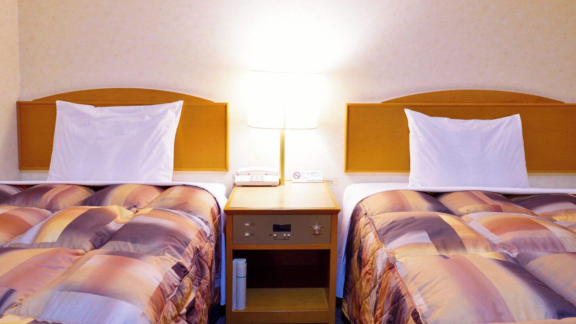 Hotellihuone, jossa on kaksi sänkyä, yöpöytä lampun kanssa, puhelin ja digitaalinen kello.