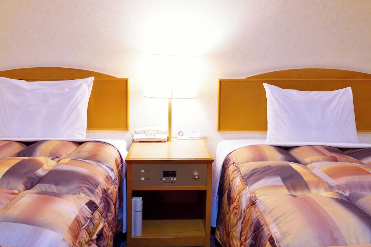 A hotel room with two beds, a bedside table with a lamp, a phone, and a digital clock.