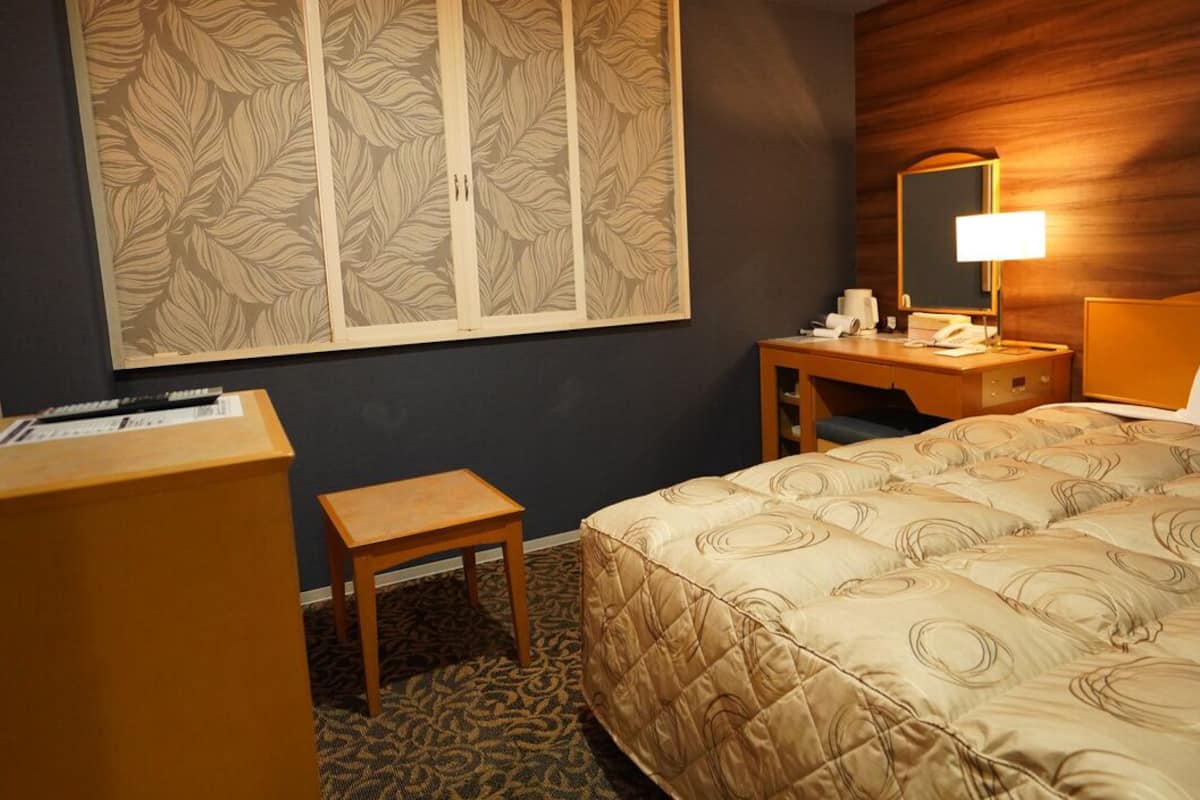 A hotel room with a bed, a desk, a chair, a small table, a mirror, and a wall with a leaf-patterned design.