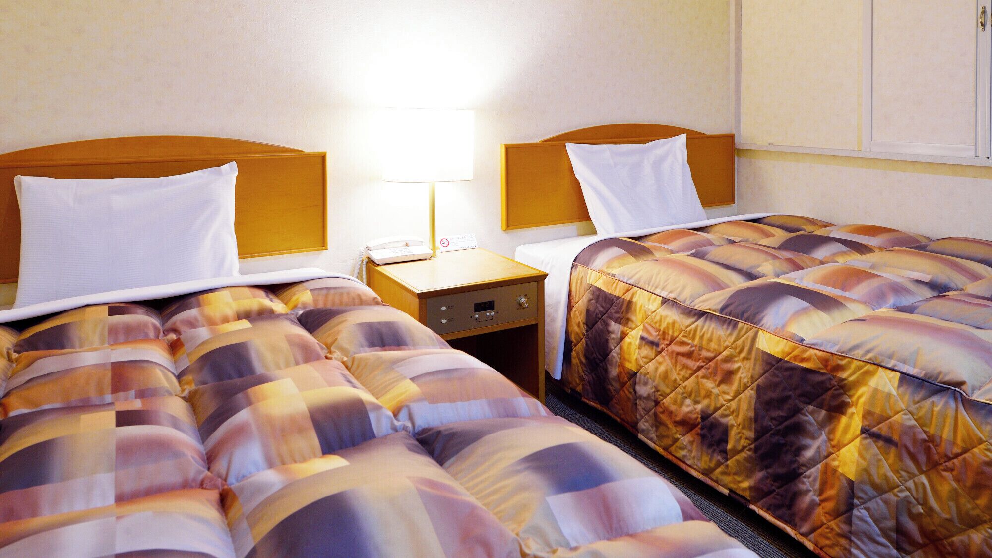Hotellihuone, jossa on kaksi sänkyä, yöpöytä lampun kanssa ja seinälle kiinnitetty kaappi.