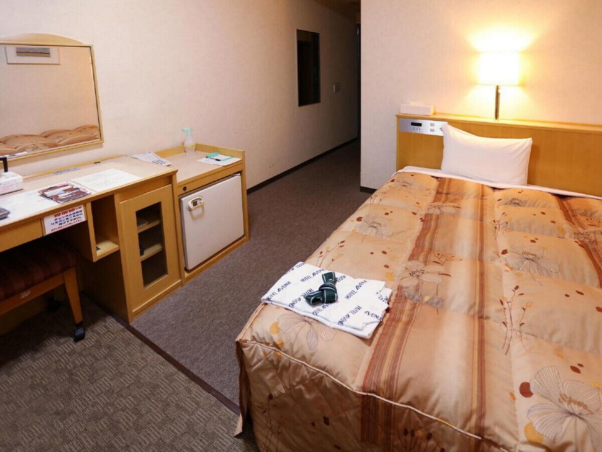 Hotellihuone, jossa on sänky, työpöytä tuolilla, pieni jääkaappi ja yöpöytä lampulla.