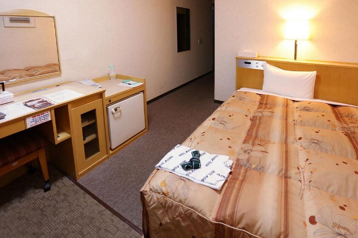A hotel room with a bed, a desk with a chair, a small refrigerator, and a nightstand with a lamp.
