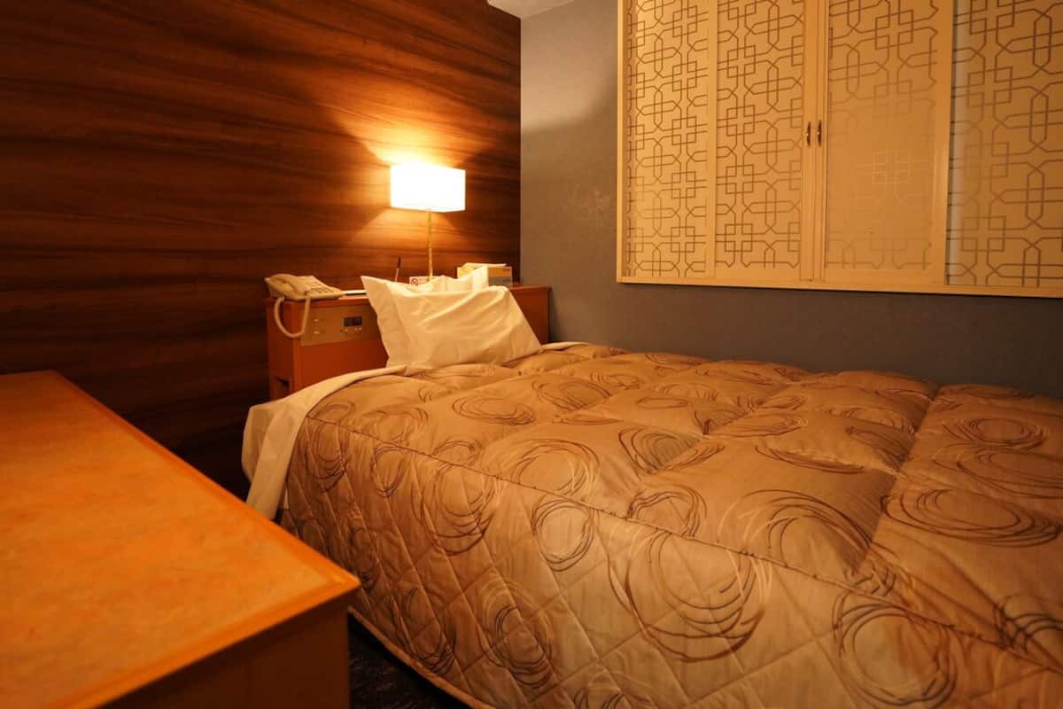 A hotel room with a wooden wall, a bed with a patterned bedspread, a bedside table with a lamp, and a wardrobe with a geometric pattern.