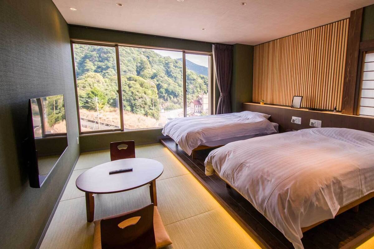 A hotel room with two beds, a TV, a small table, and a view of a forest.