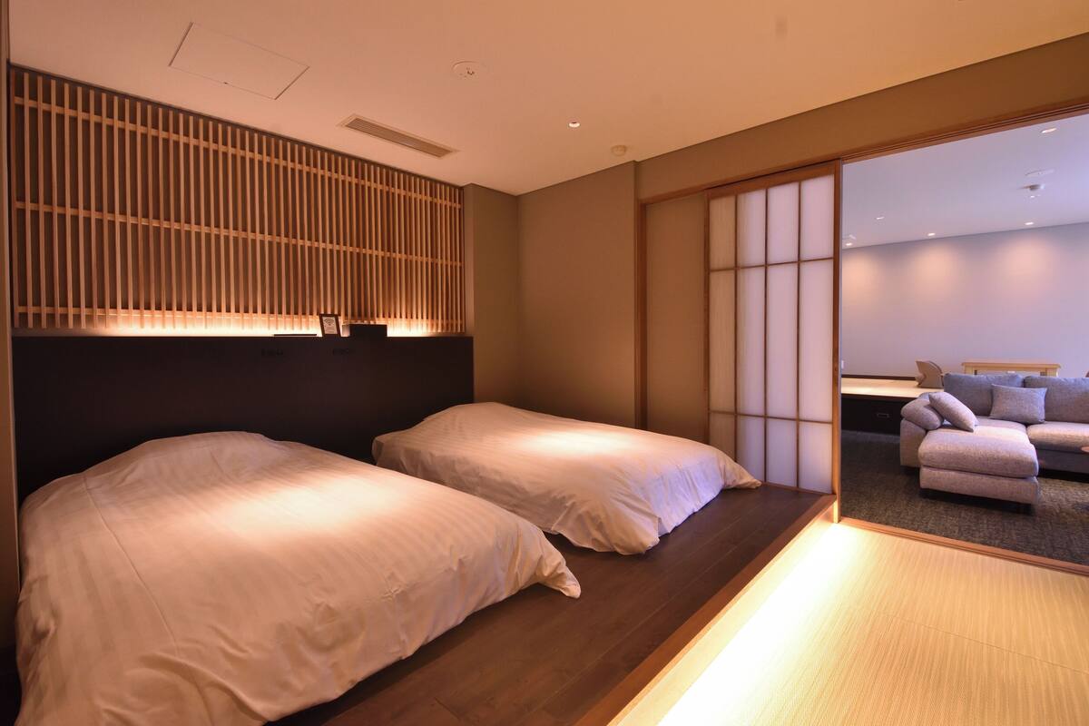 A hotel room with two beds, a wooden headboard, a sliding shoji screen, and a sofa in the adjacent living area.