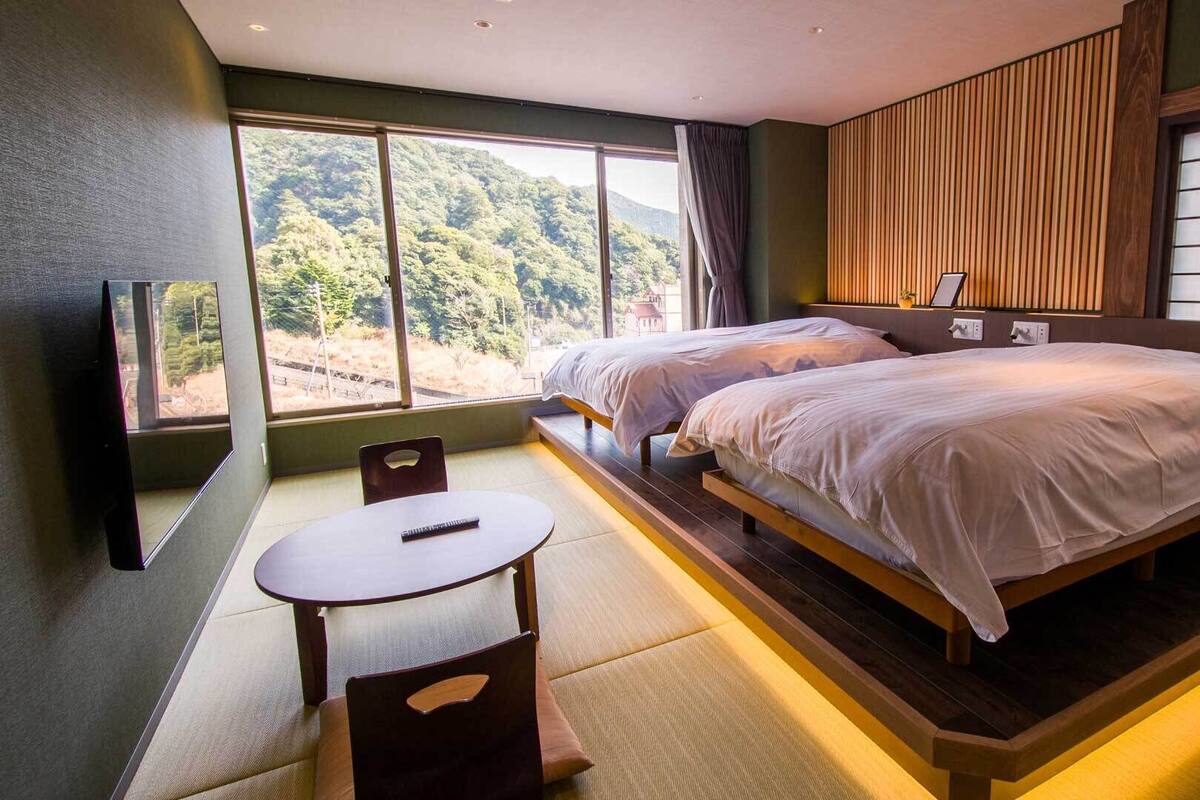 A bedroom with two beds, a TV, a table, and a view of a forest.