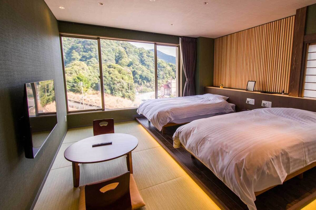 A hotel room with two beds, a TV, a small table, and a view of a forest.