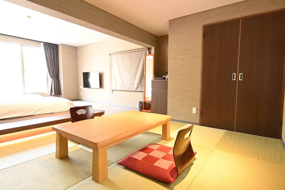 A hotel room with a bed, a wooden table, a chair, a TV, a wardrobe, and tatami mats.