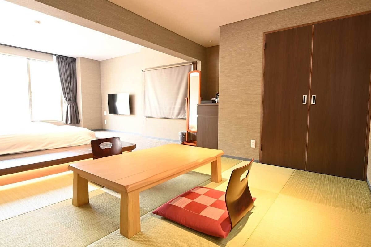 Una habitación de hotel con cama, una mesa de madera, una silla, televisor, armario y esteras de tatami.