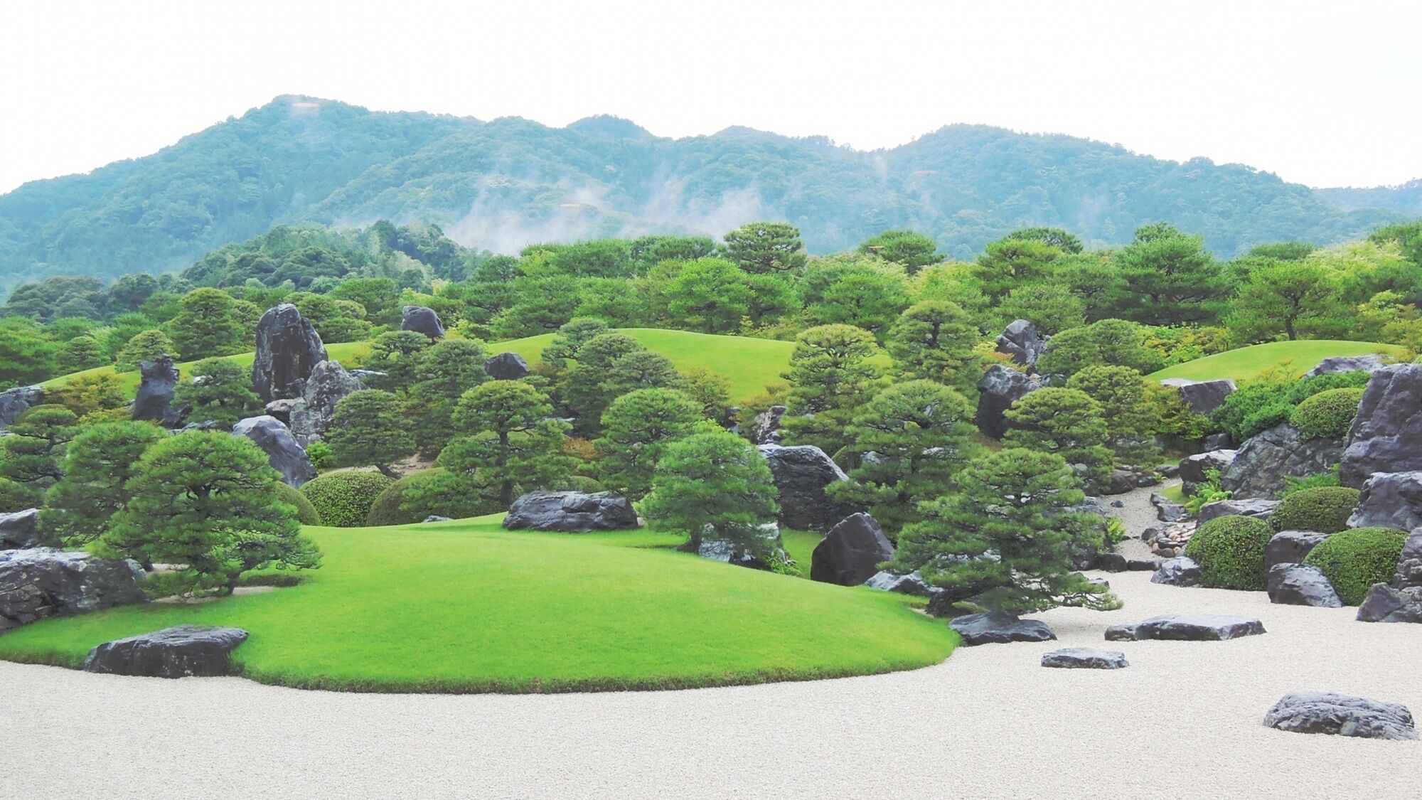 A serene landscape with a well-maintained garden, featuring neatly trimmed grass, various trees, and large rocks.