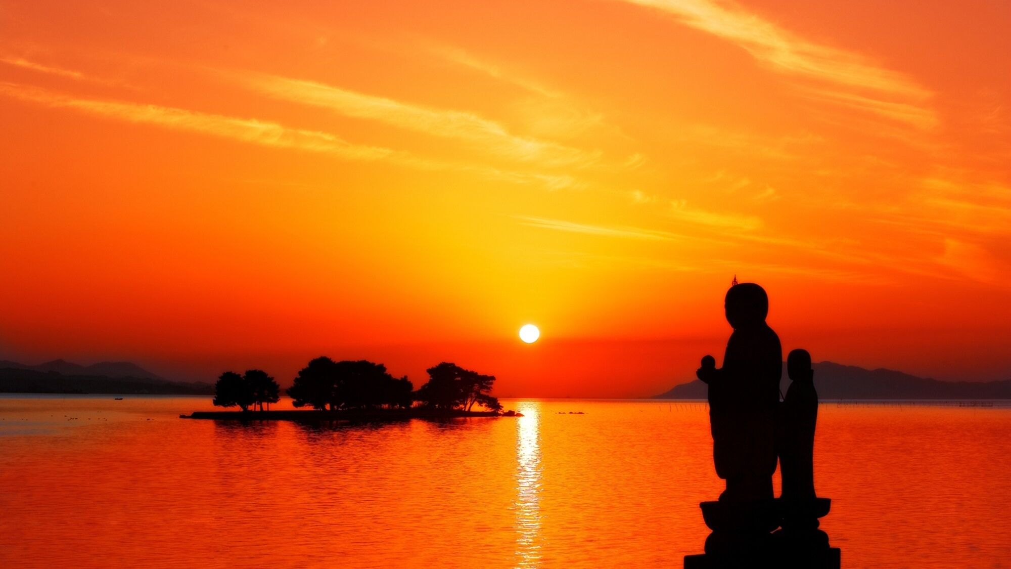A sunset over a calm body of water with a silhouette of a statue and a small island with trees.