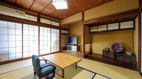 A traditional Japanese room with tatami flooring, a low table, a blue patterned chair, a television, and a shelf with decorative items.