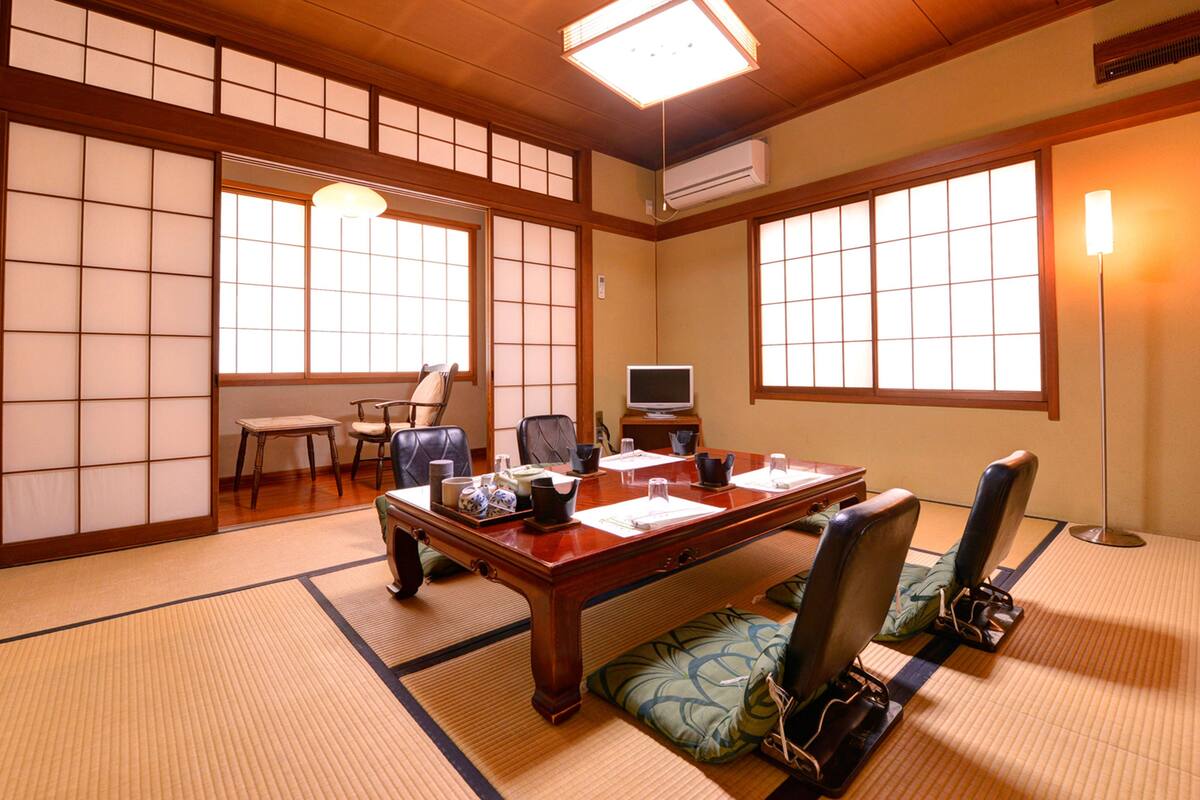 A traditional Japanese-style room with tatami flooring, a wooden table, chairs, a television, and a large window providing natural light.
