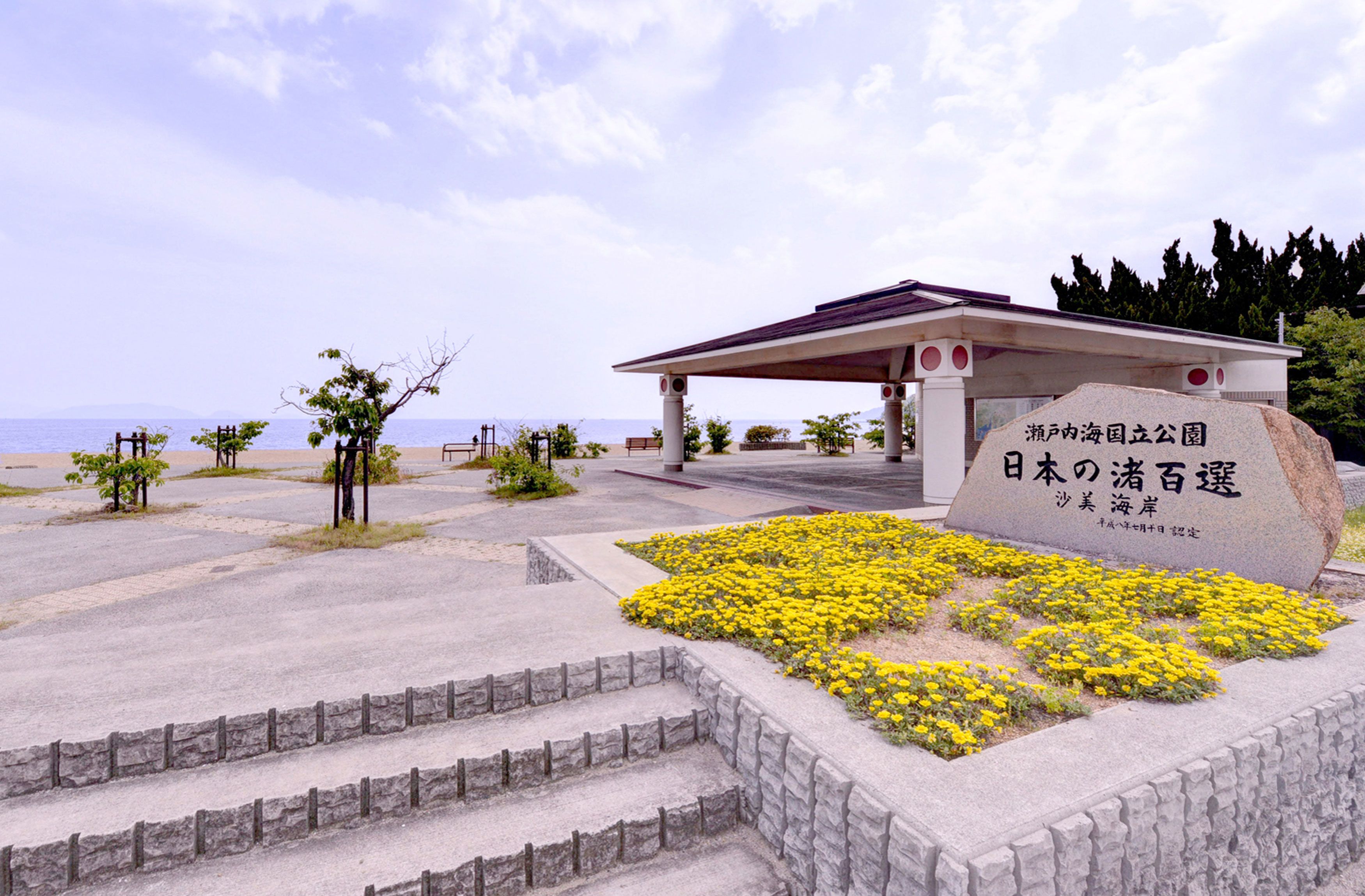 Um monumento de pedra com texto em japonês, um pavilhão coberto e uma área ajardinada com flores amarelas e degraus que levam até ele.