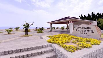 Um monumento de pedra com texto em japonês, um pavilhão coberto e uma área ajardinada com flores amarelas e degraus que levam até ele.