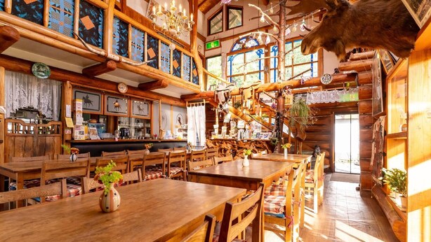 A rustic wooden interior with high ceilings, exposed beams, and a variety of hanging decorations. Tables and chairs are arranged for dining, and a large animal head is mounted on the wall. - Hope Lodge (Chino)