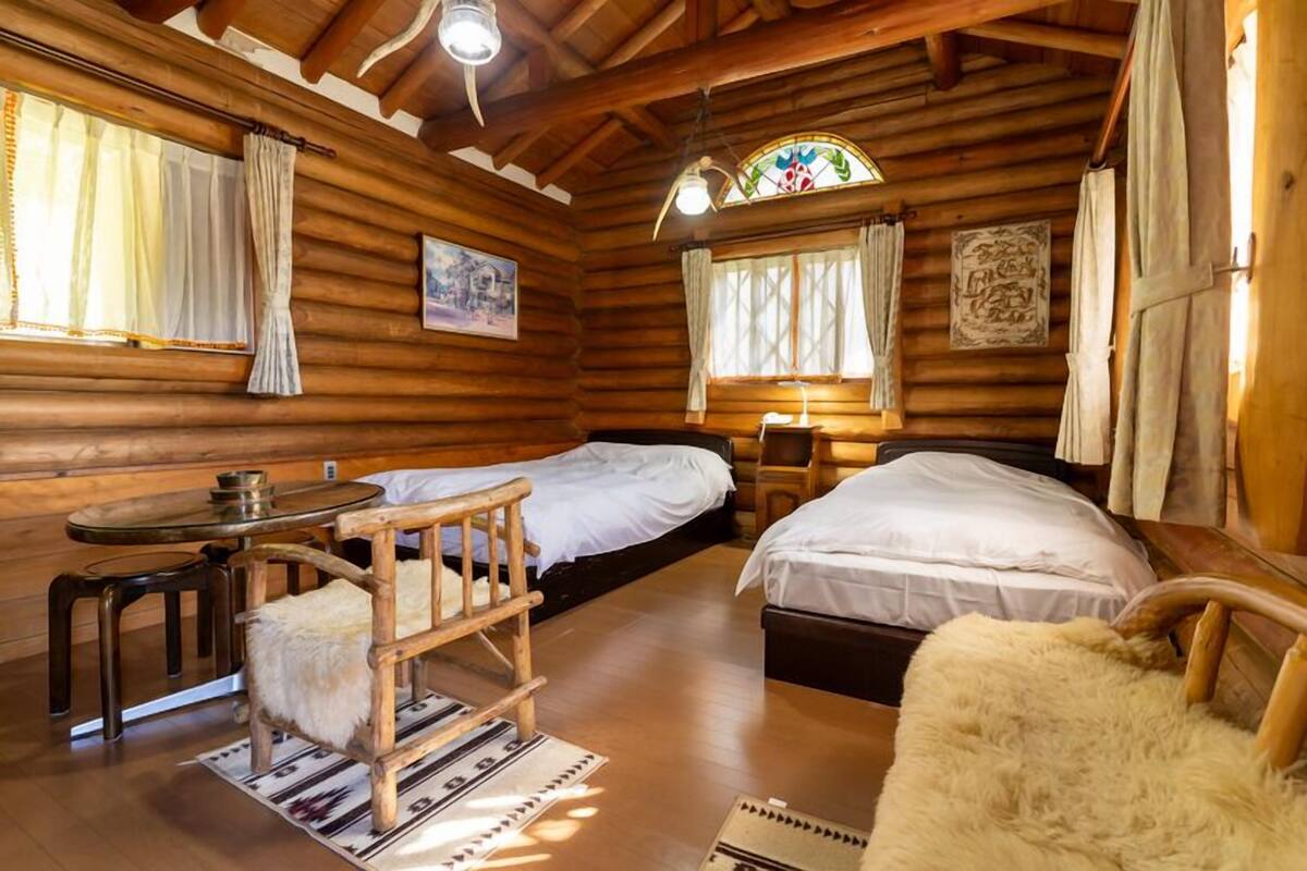 A log cabin interior with two beds, a dining area with a table and chairs, and a window with curtains.