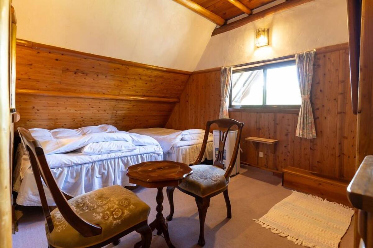 A room with two beds, a wooden ceiling, a small table, two chairs, and a window with curtains.