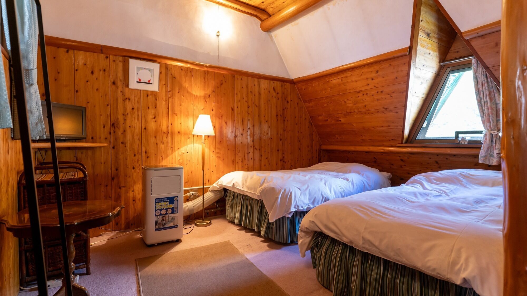 A cozy cabin room with wooden walls, two beds, a desk with a lamp, and a television.