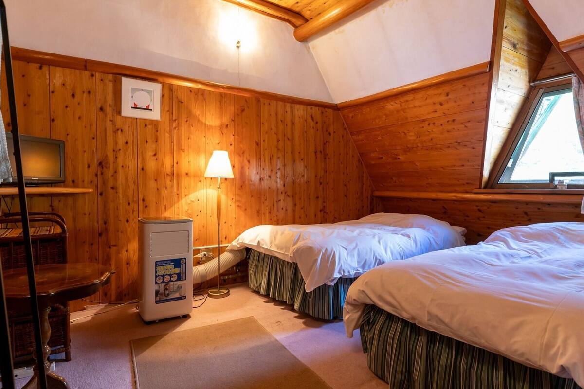 A cozy cabin room with wooden walls, two beds, a desk with a lamp, and a television.
