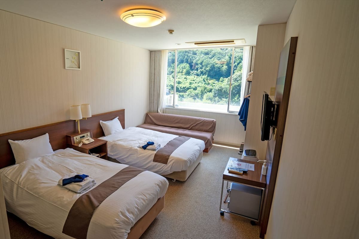A hotel room with two beds, a bedside table with a lamp, a window with a view of greenery, and a small desk with a telephone.