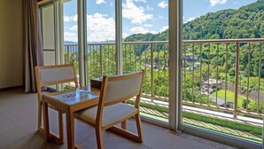 Una habitación con balcón, una mesa de madera, dos sillas y vistas a un paisaje verde con árboles y edificios.