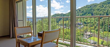 Una habitación con balcón, una mesa de madera, dos sillas y vistas a un paisaje verde con árboles y edificios.