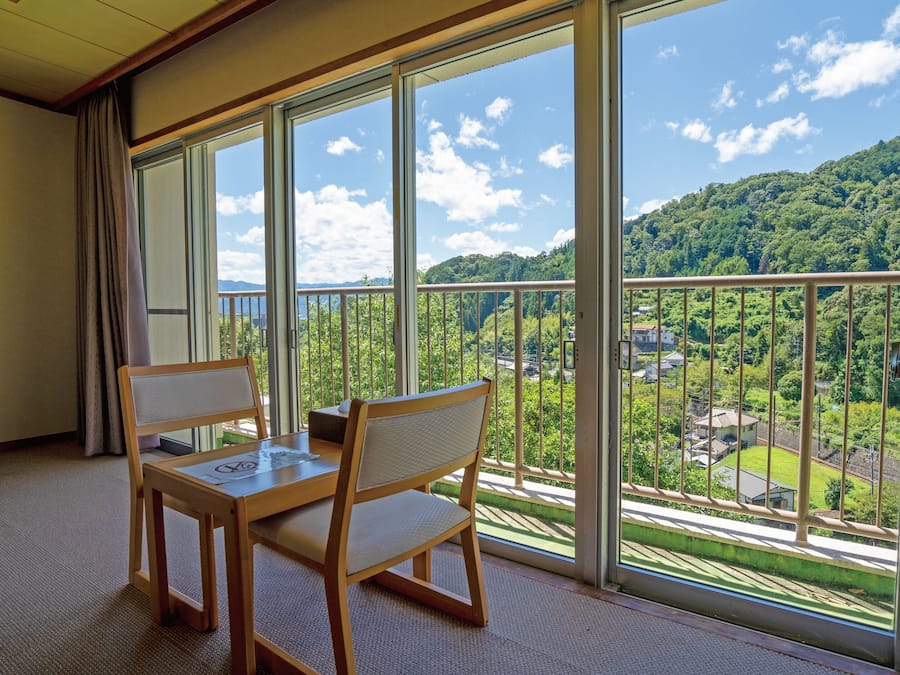 A room with a balcony, a wooden table, two chairs, and a view of a green landscape with trees and buildings.
