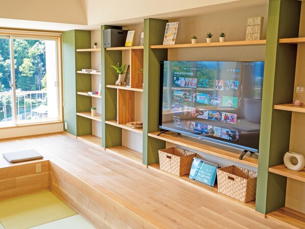 Una sala de estar moderna con un televisor de pantalla plana grande mostrando la interfaz de Netflix, estanterías de madera con diversos objetos, una ventana con vista a árboles y un tatami en el suelo.