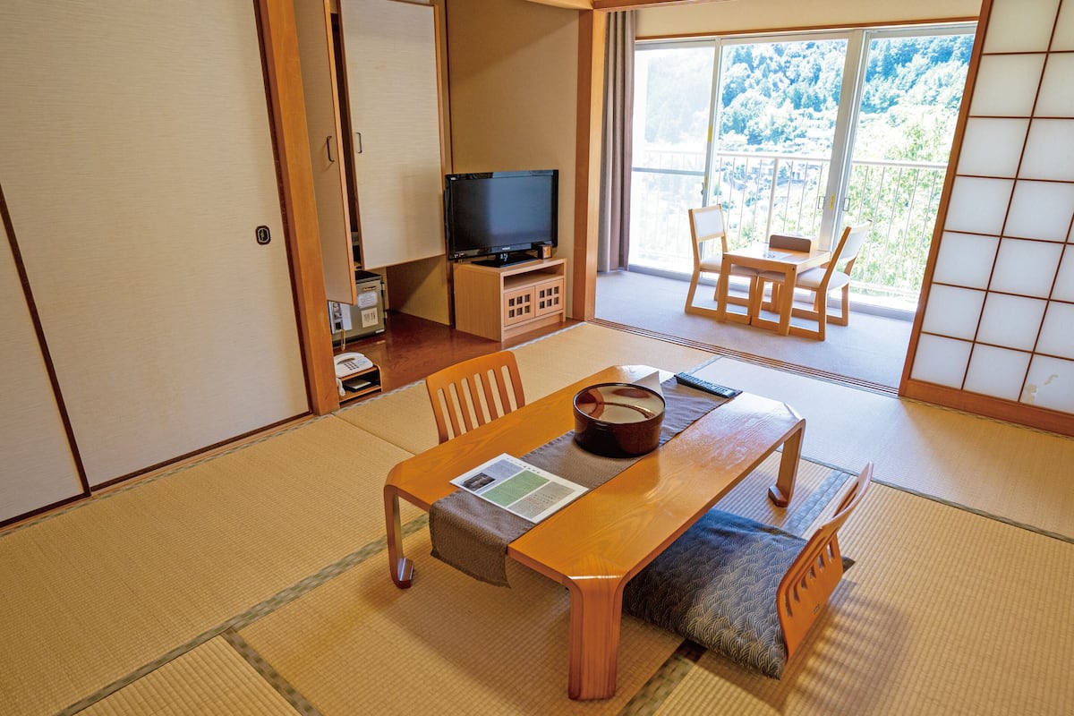 A traditional Japanese room with tatami flooring, a wooden table with a tea set, a television, and a sliding door leading to a balcony with a view of trees.