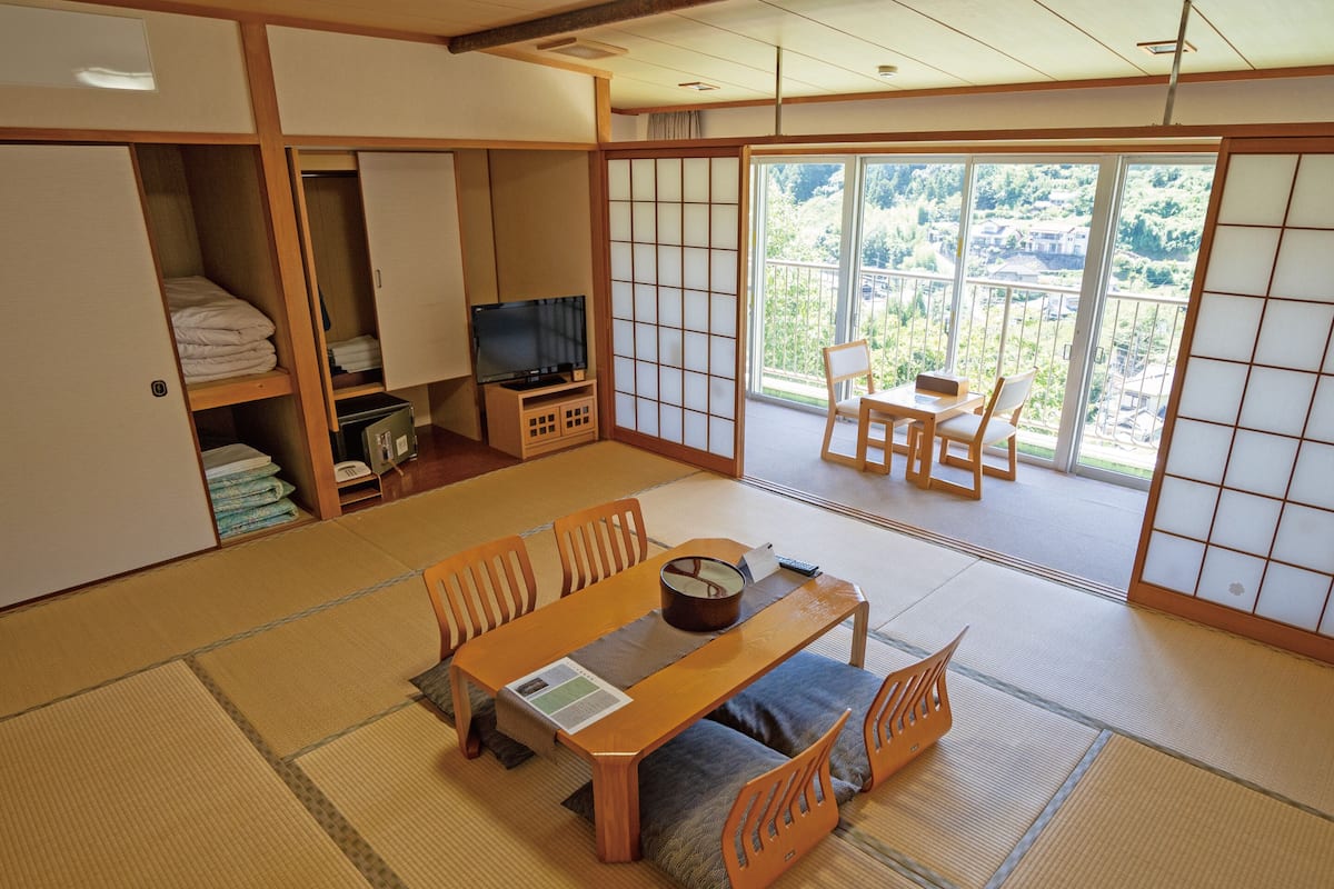A traditional Japanese-style room with tatami flooring, a low table with cushions, a television, and sliding doors leading to a balcony with a view of a residential area.