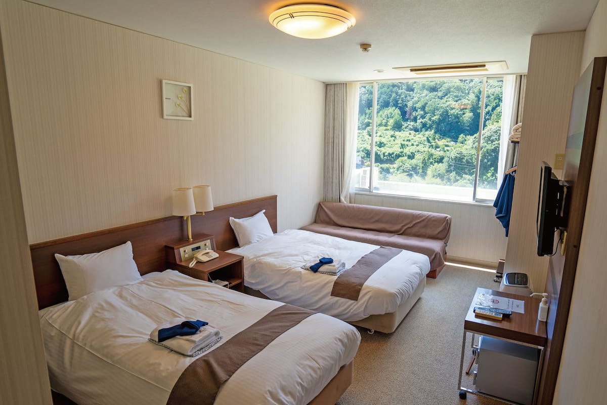 A hotel room with two beds, a bedside table with a phone, a window with a view of greenery, and a desk with a computer and a bottle.