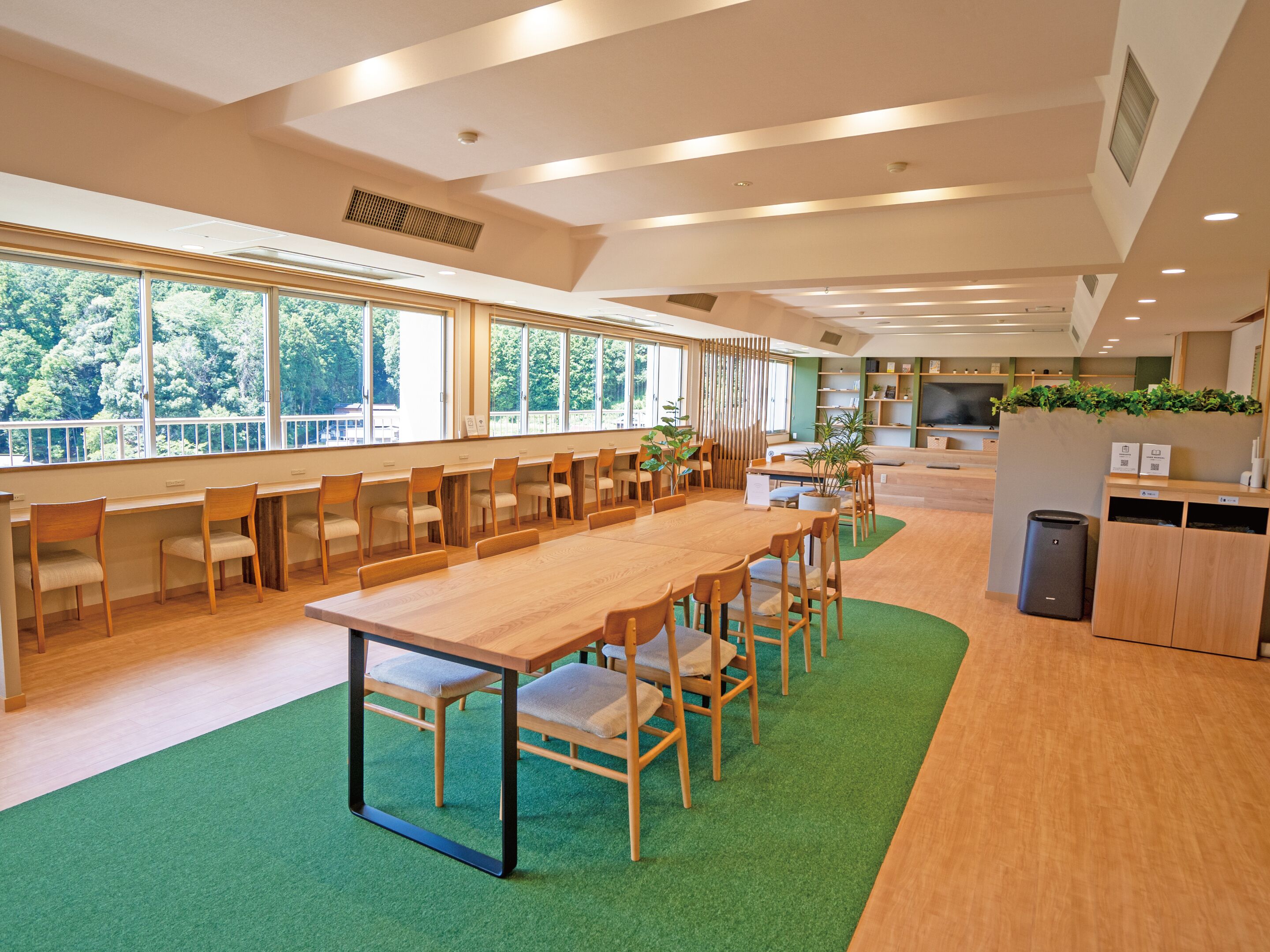 A spacious indoor area with long wooden tables and chairs, large windows offering a view of greenery outside, and a green carpet on the floor.